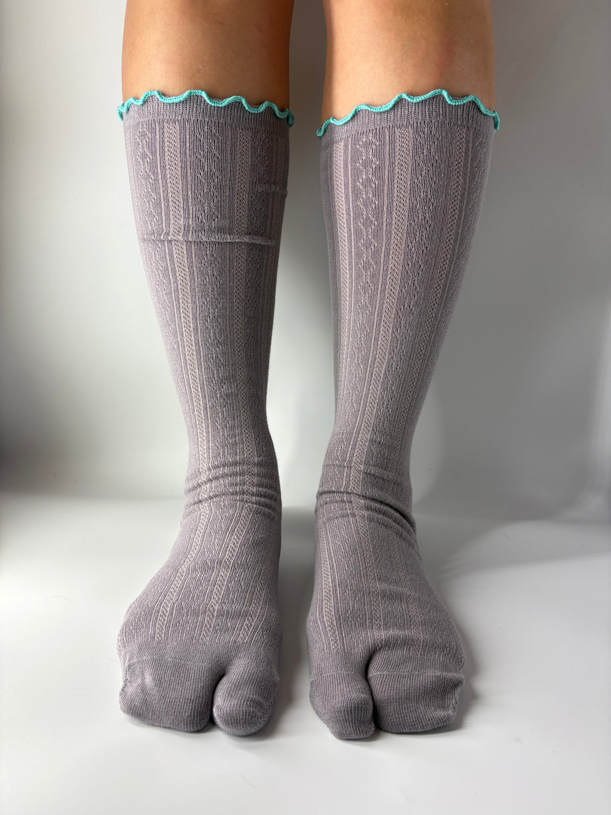 A pair of gray ribbed split toe socks with a turquoise scalloped trim at the top, pointelle knit shown on a person's feet against a plain light gray background.