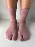 A pair of ribbed, scalloped-edge crew socks in a dusty rose pink color, shown on a person's feet in a traditional five-toe split sock style against a neutral light gray background.