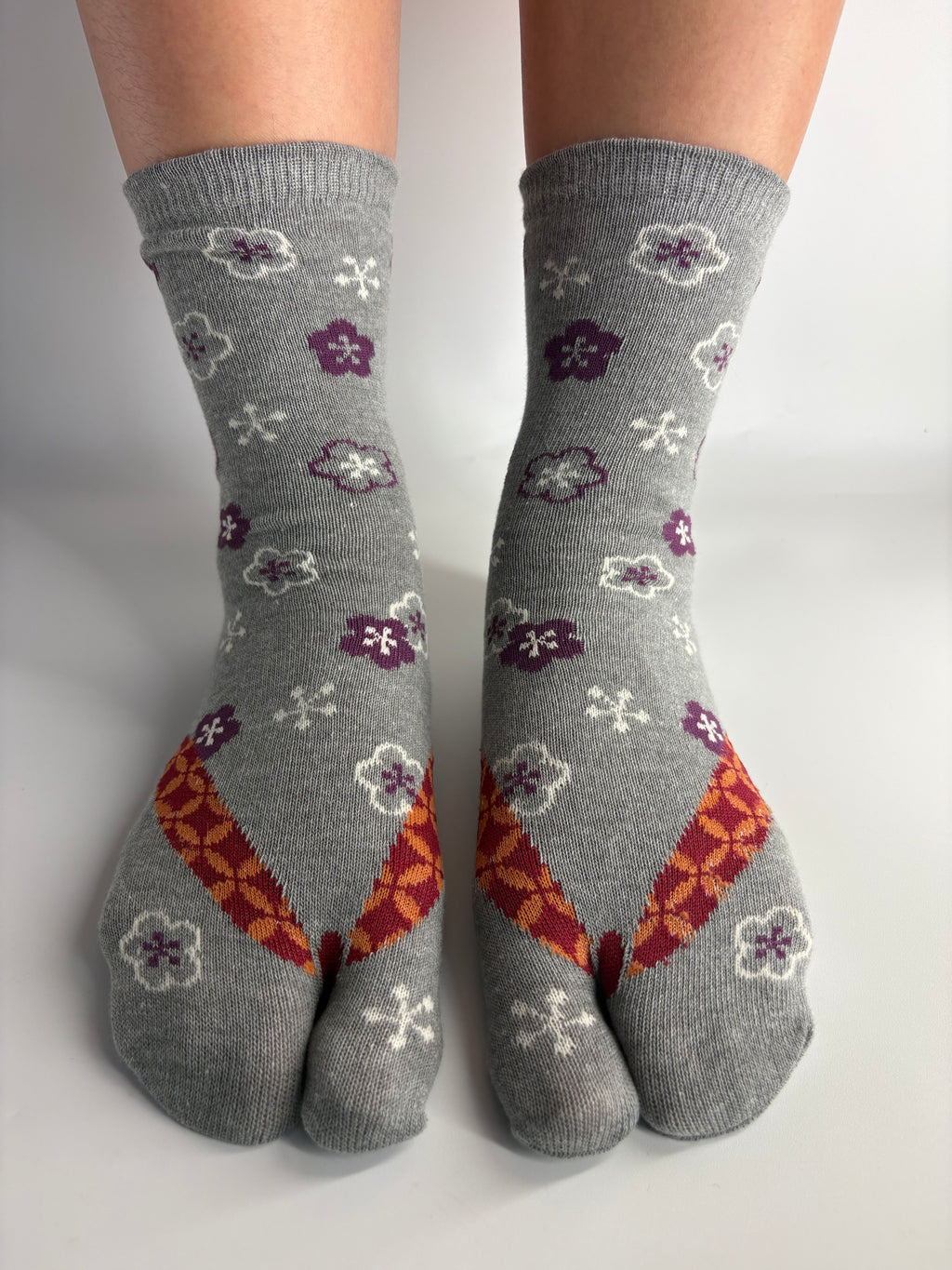 A pair of grey split-toe socks with a pattern of purple and white flowers and orange geometric accents, shown on feet against a plain white background.