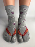 A pair of grey split-toe socks with a pattern of purple and white flowers and orange geometric accents, shown on feet against a plain white background.