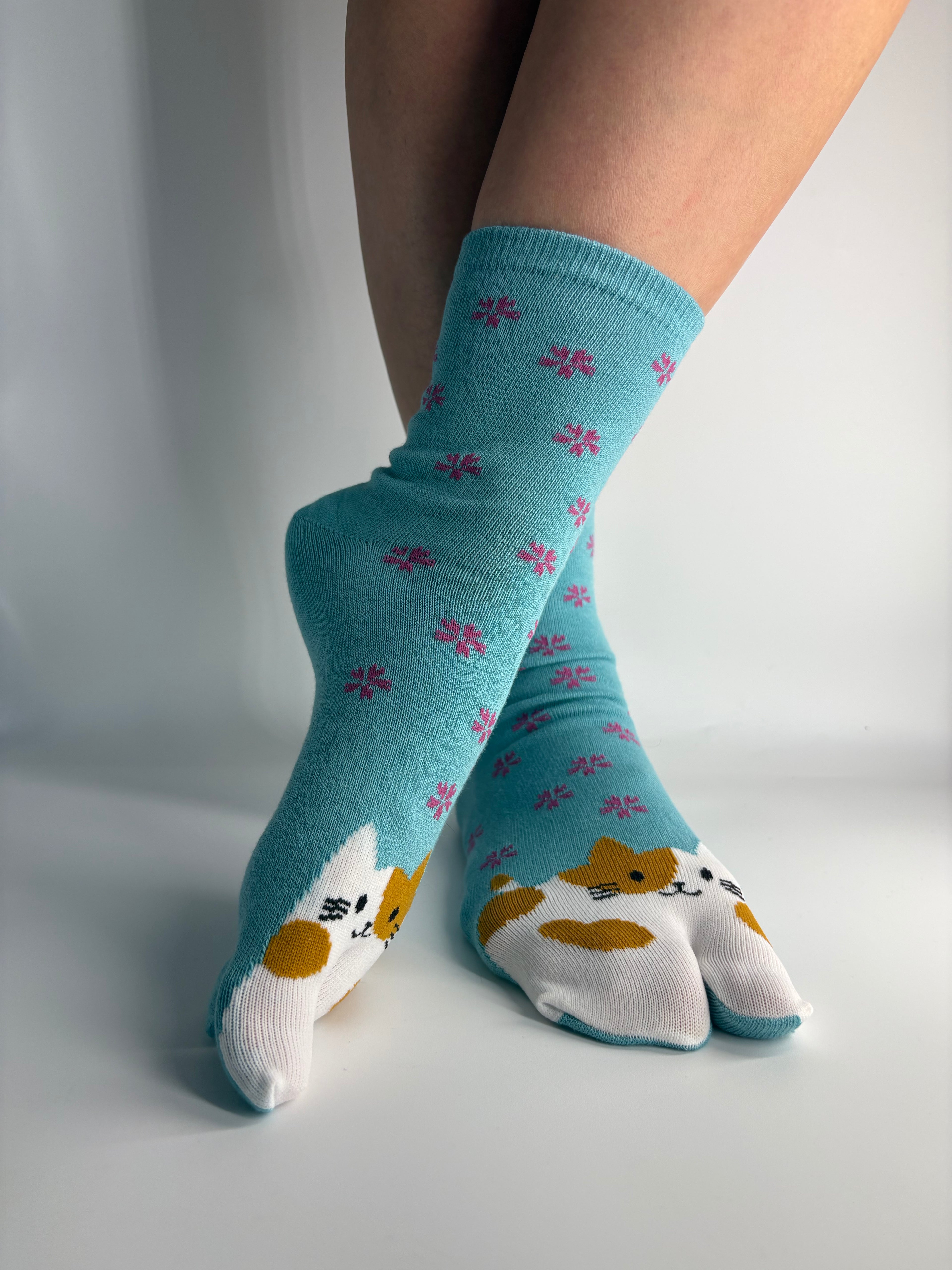 A pair of turquoise tabi split toe socks with a white cat face pattern and small pink floral motifs, shown on feet against a plain light background.
