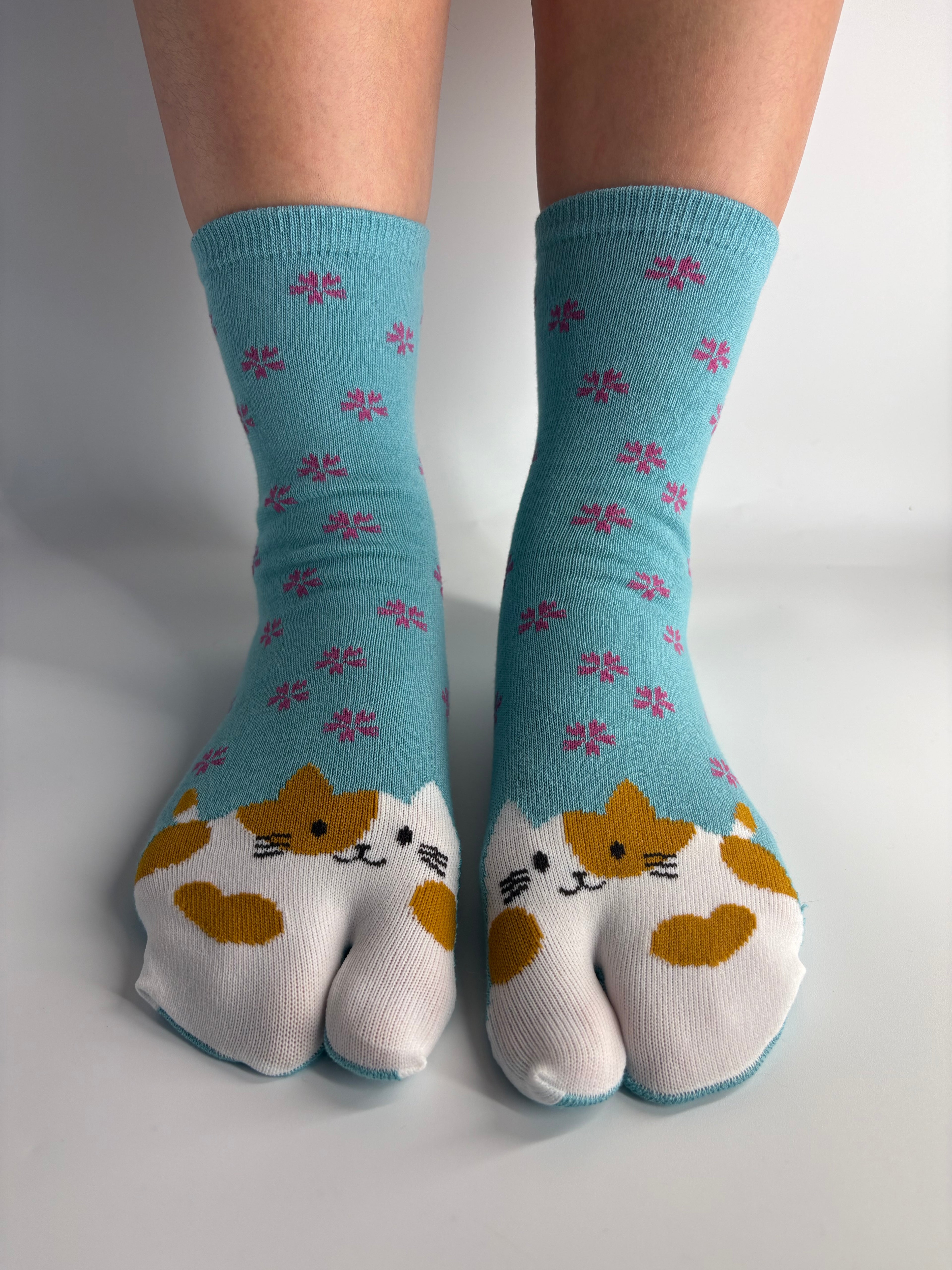 A pair of turquoise tabi split toe socks with a white cat face pattern and small pink floral motifs, shown on feet against a plain light background.