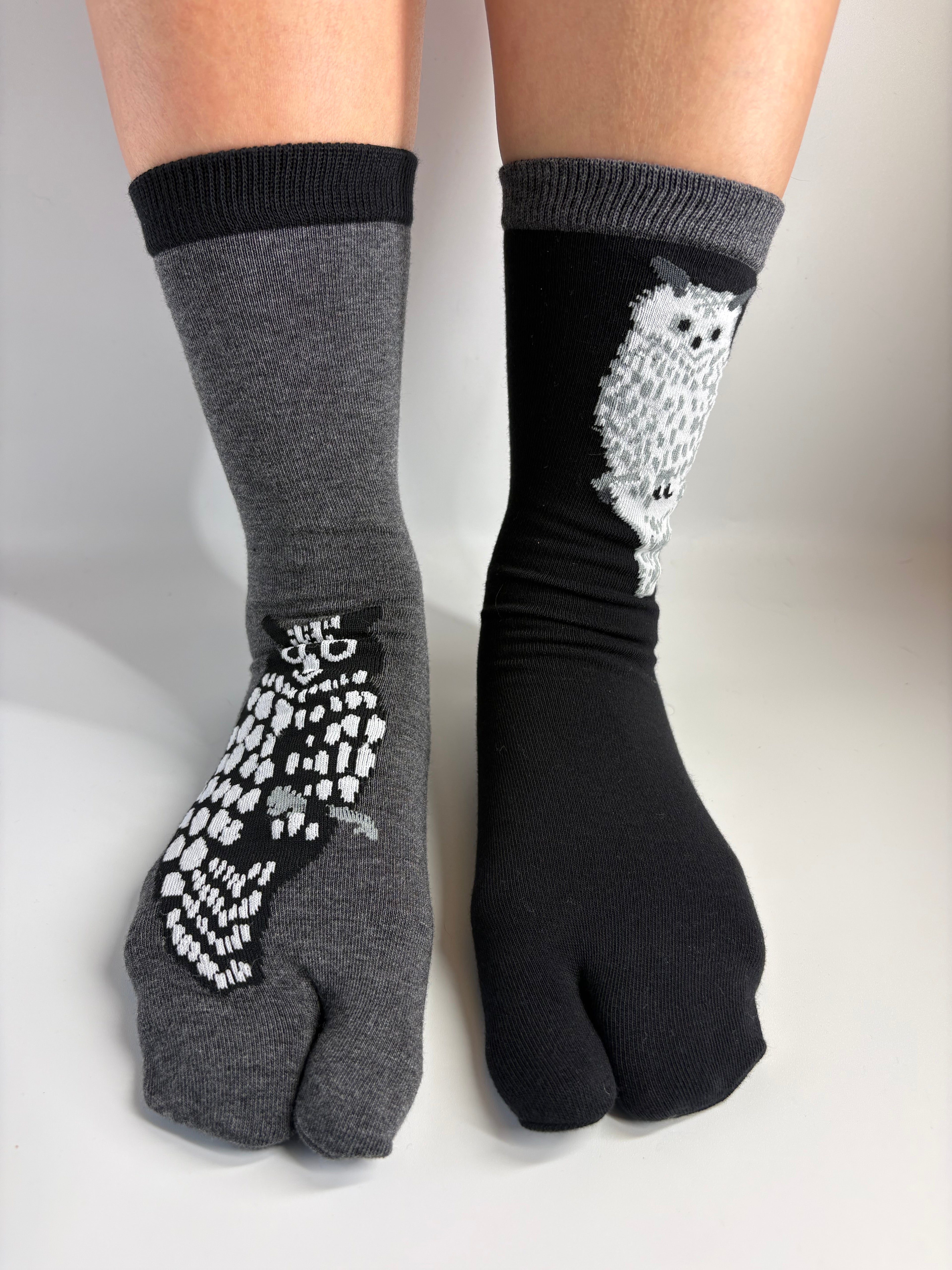 A pair of split-toe socks shown on feet, with one sock in heather gray featuring a white owl print and one sock in black featuring a white owl design.