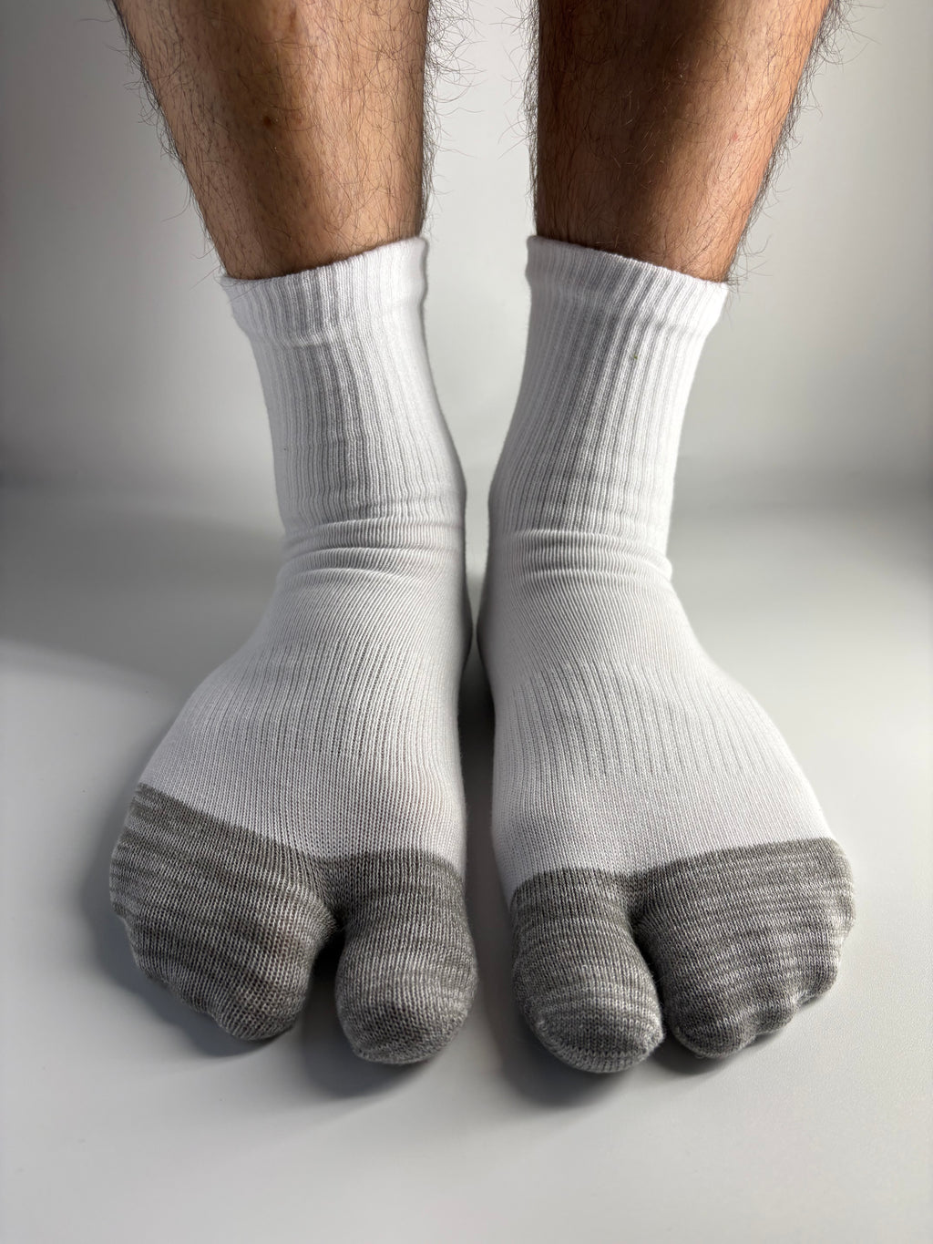 A close-up of a person's feet wearing white ribbed socks with gray toe caps, resting against a light gray wall.