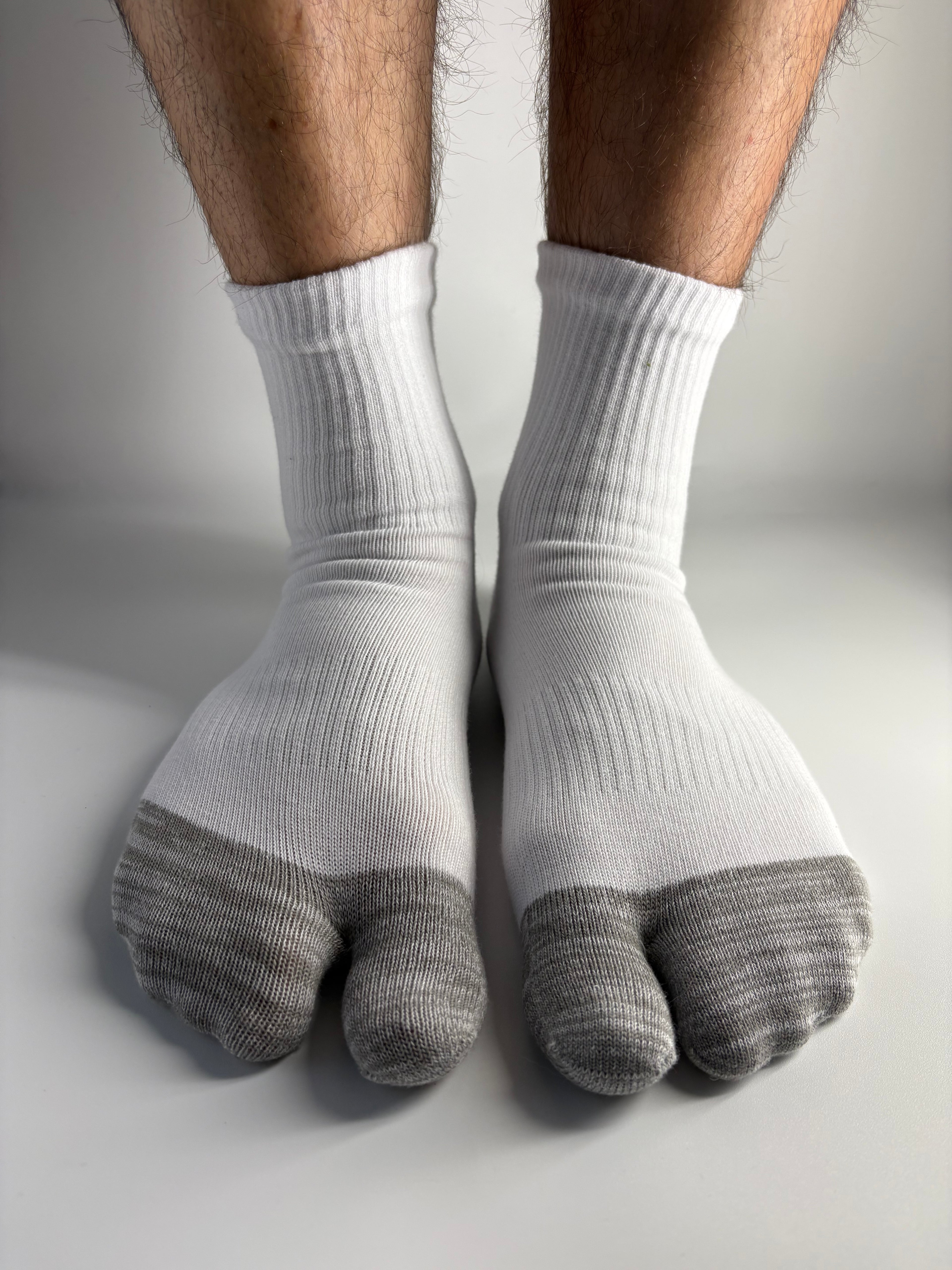 A close-up of a person's feet wearing white ribbed socks with gray toe caps, resting against a light gray wall.