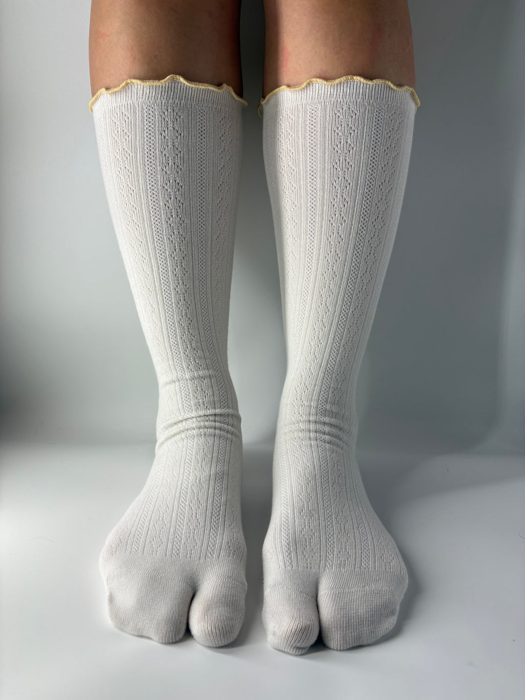 A pair of off-white ribbed knit tabi split toe socks worn on a person's feet, featuring a scalloped yellow trim at the top.