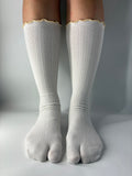 A pair of off-white ribbed knit tabi split toe socks worn on a person's feet, featuring a scalloped yellow trim at the top.