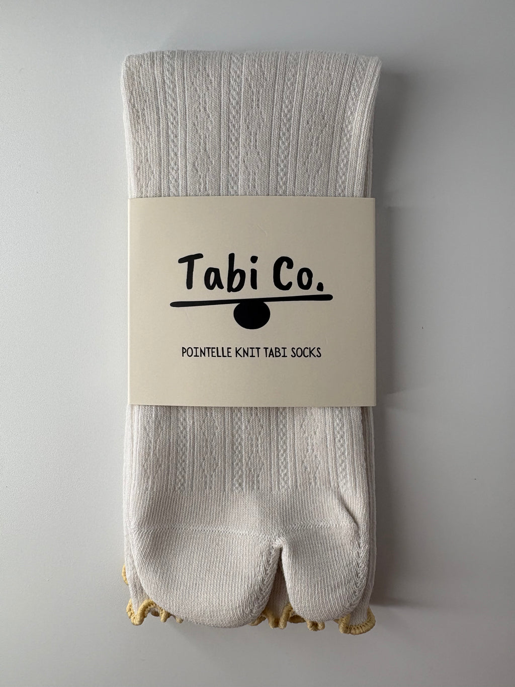 A pair of off-white ribbed tabi socks folded with a beige paper band label that reads 'Tabi Co. POINTELLE KNIT TABI SOCKS' on a light gray background.